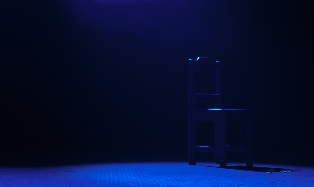 A solitary wooden chair is lit by a dim blue spotlight in an otherwise dark room, casting a shadow. The scene evokes a sense of loneliness and introspection.