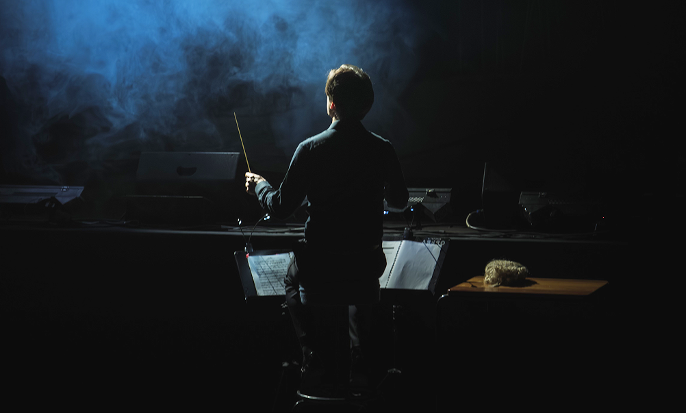 A conductor stands in front of a music stand, facing a dark stage with smoke or fog and faint blue lighting.