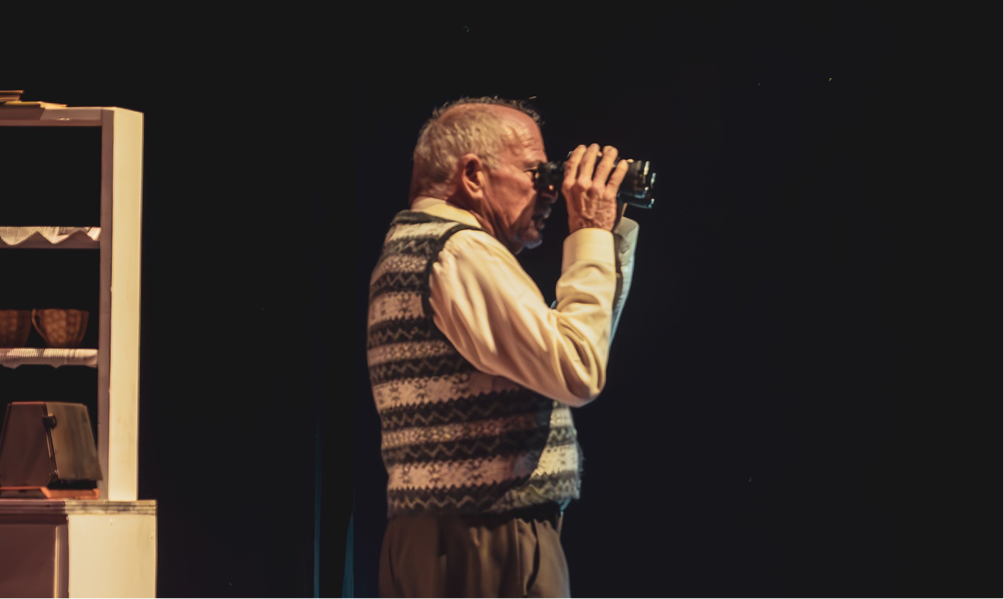 Elderly man in a patterned sweater vest gazes through binoculars, standing focused on a dimly lit stage with a white cabinet partially visible.