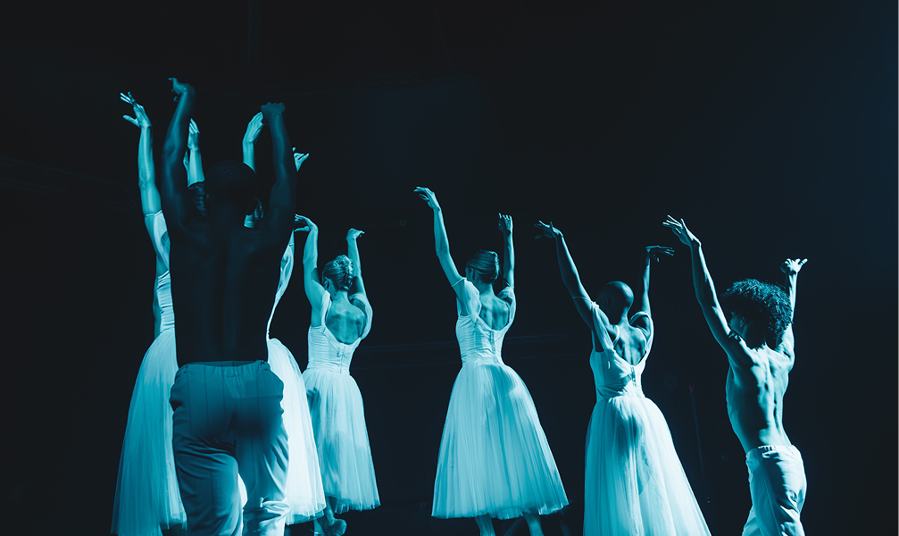 Six ballet dancers, three male and three female, perform on stage with their arms raised, wearing light-colored costumes under blue stage lighting.